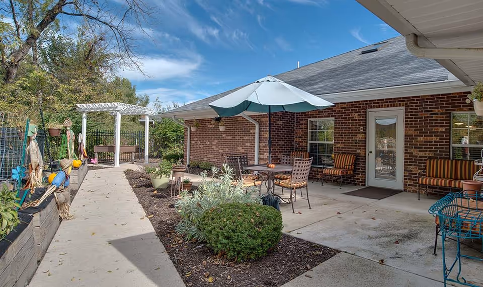 Outdoor patio area at Chestnut Glen Senior Living featuring a concrete walkway, garden beds with plants and decorations, a white pergola, and a seating area with a table, chairs, and an umbrella next to a brick building under a blue sky with some clouds.