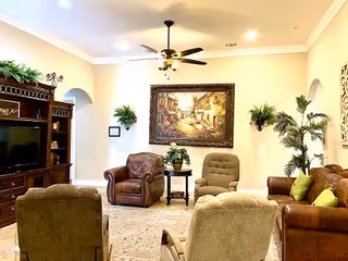 A cozy living room with a ceiling fan and light fixture, featuring a large framed painting on the wall. The room has several upholstered chairs and a leather sofa with green pillows arranged around a small wooden side table with a plant. There is a dark wooden entertainment center with a TV and decorative greenery on top.
