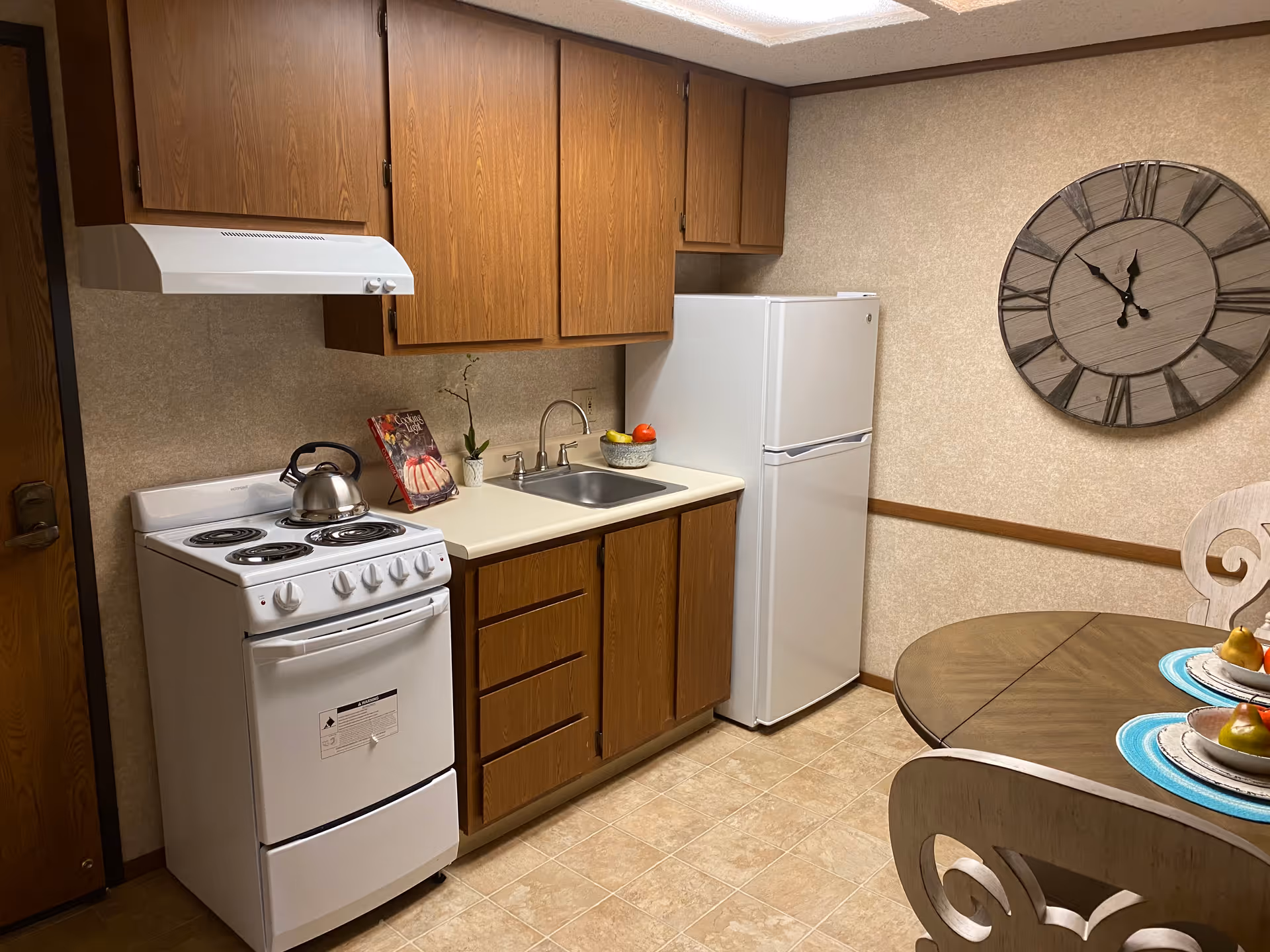 Small kitchenette with a white stove, refrigerator, sink, wooden cabinets, round dining table and large wall clock.
