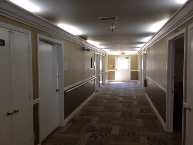 Carpeted corridor inside a senior living facility with doors along both sides and an exit door at the far end.