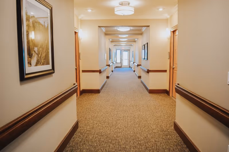 A well-lit, carpeted hallway in a senior living facility with beige walls, wooden handrails, framed artwork on the walls, and multiple doors on either side leading to rooms. The hallway extends to a glass door at the far end.