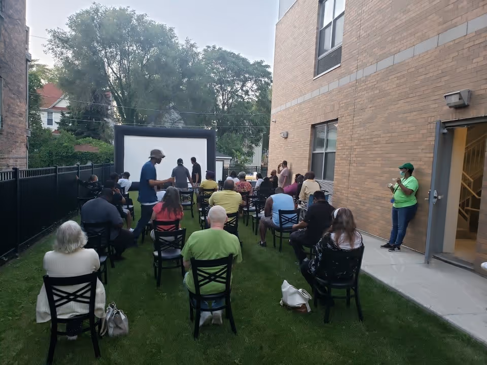 Residents and guests sit on chairs in a grassy courtyard watching a large inflatable outdoor movie screen beside a brick building.