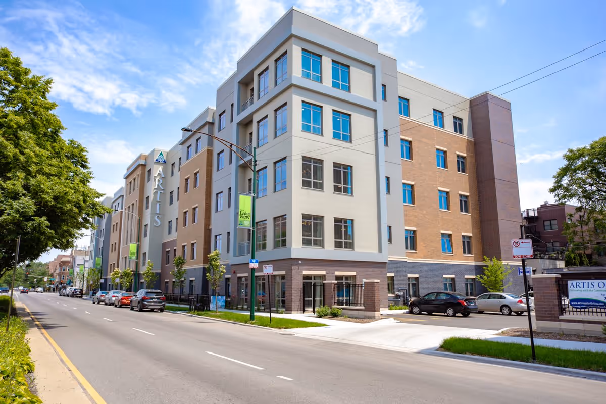Exterior view of a modern multi-story senior living facility named Artis Senior Living of Lakeview, with a clear blue sky, trees along the street, parked cars, and a sidewalk.