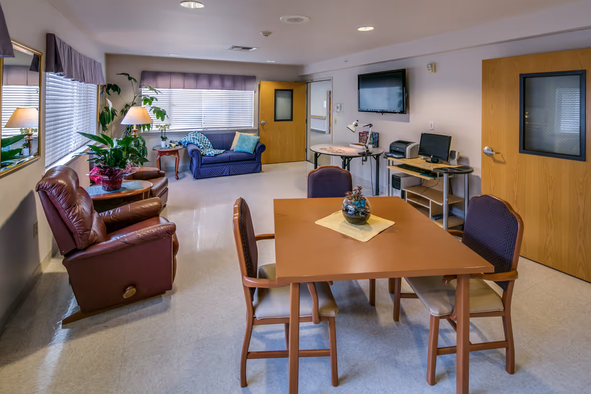 A well-lit common area in a senior living facility featuring a square wooden table with four chairs, a small plant centerpiece, two brown leather recliners, a blue loveseat with cushions, a side table with a lamp and plants, a wall-mounted TV, a computer desk with a printer, and two wooden doors with windows.