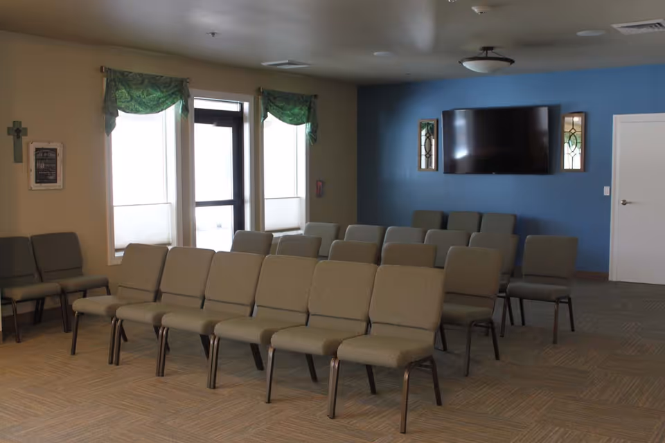 A room with multiple rows of beige cushioned chairs arranged facing a wall-mounted flat screen TV. The walls are painted beige and blue, with two windows covered by green valances and a door in the background. The room appears to be a meeting or activity space in a senior living facility.
