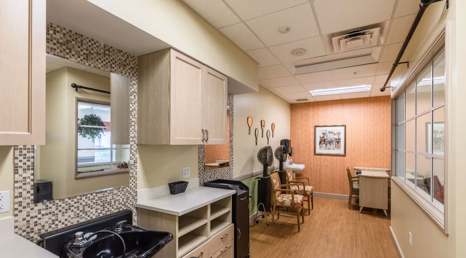 Interior view of a salon area in a senior living facility featuring two vintage hair dryers with chairs underneath, a black sink with a countertop and cabinets, a large mirror with mosaic tile border, a small sink on the far wall, decorative wall art shaped like hairbrushes, and a framed picture on an orange textured wall. There is a window with multiple panes on the right side.