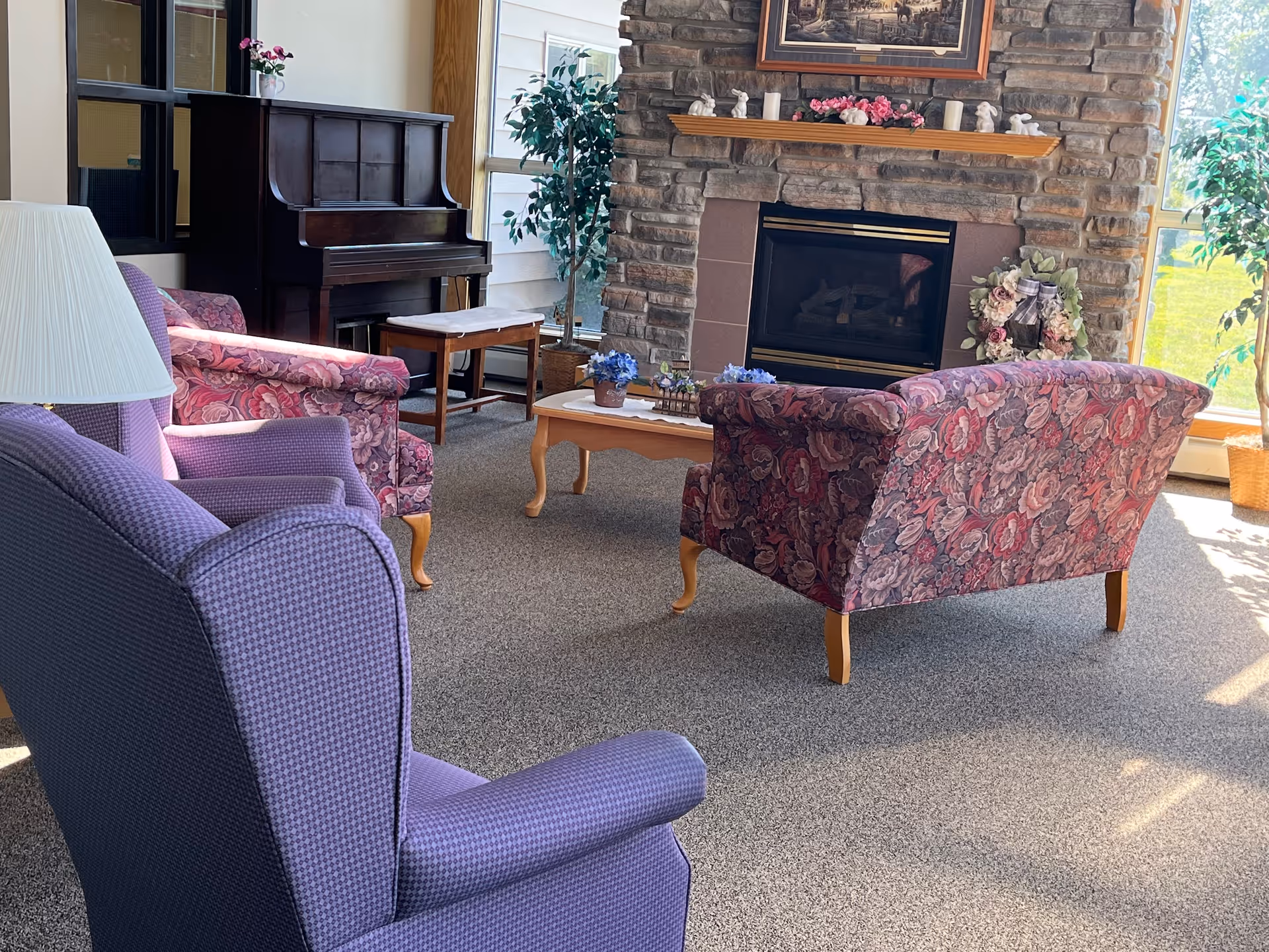 A cozy living room area with a stone fireplace decorated with small figurines and flowers on the mantel. There are floral patterned armchairs and a purple armchair arranged around a wooden coffee table with flower pots. A piano with a bench is placed near the window, and there are large windows letting in natural light with green plants nearby.