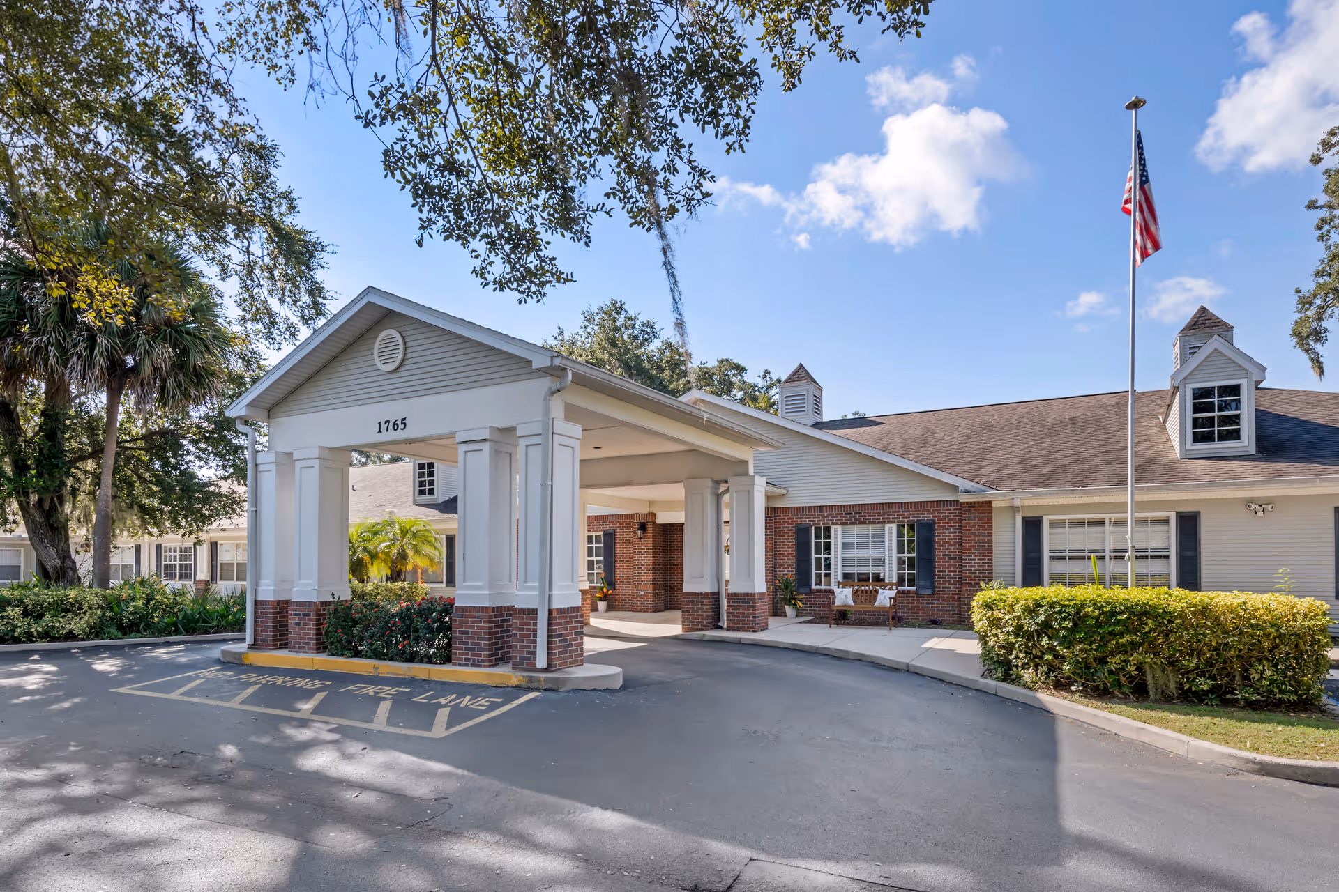 Exterior view of a senior living facility with a covered entrance supported by white columns and brick accents. The building has a beige and brick facade with windows and a flagpole flying the American flag in front. Trees and bushes surround the driveway and entrance area under a partly cloudy sky.