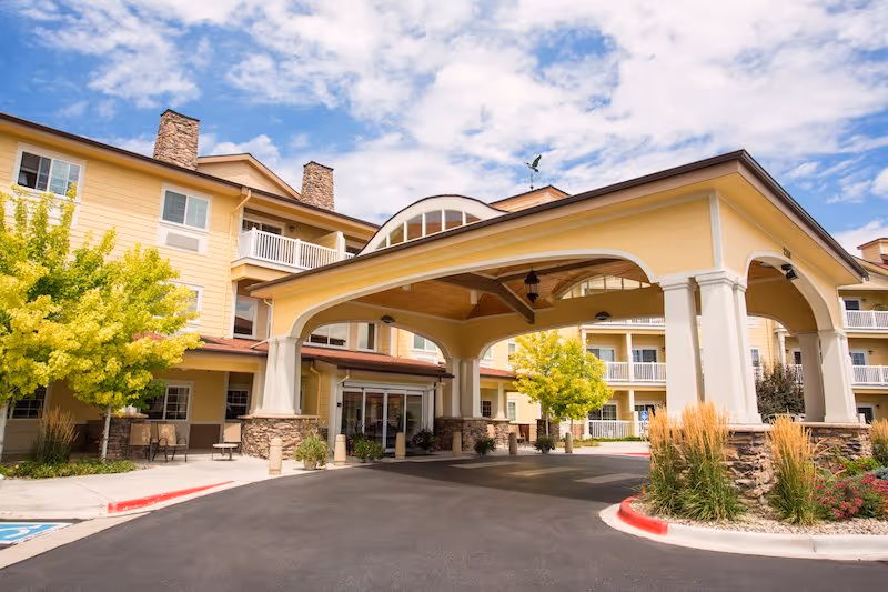 Exterior view of Rigden Farm Senior Living facility showing a large covered entrance with stone pillars and yellow siding. The building has multiple floors with balconies and windows, surrounded by landscaped greenery and a clear blue sky with some clouds.