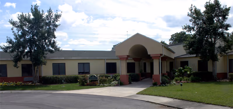 Exterior front view of a single-story rehabilitation and healing center building with a covered entrance, surrounded by green grass, shrubs, and trees under a partly cloudy sky.