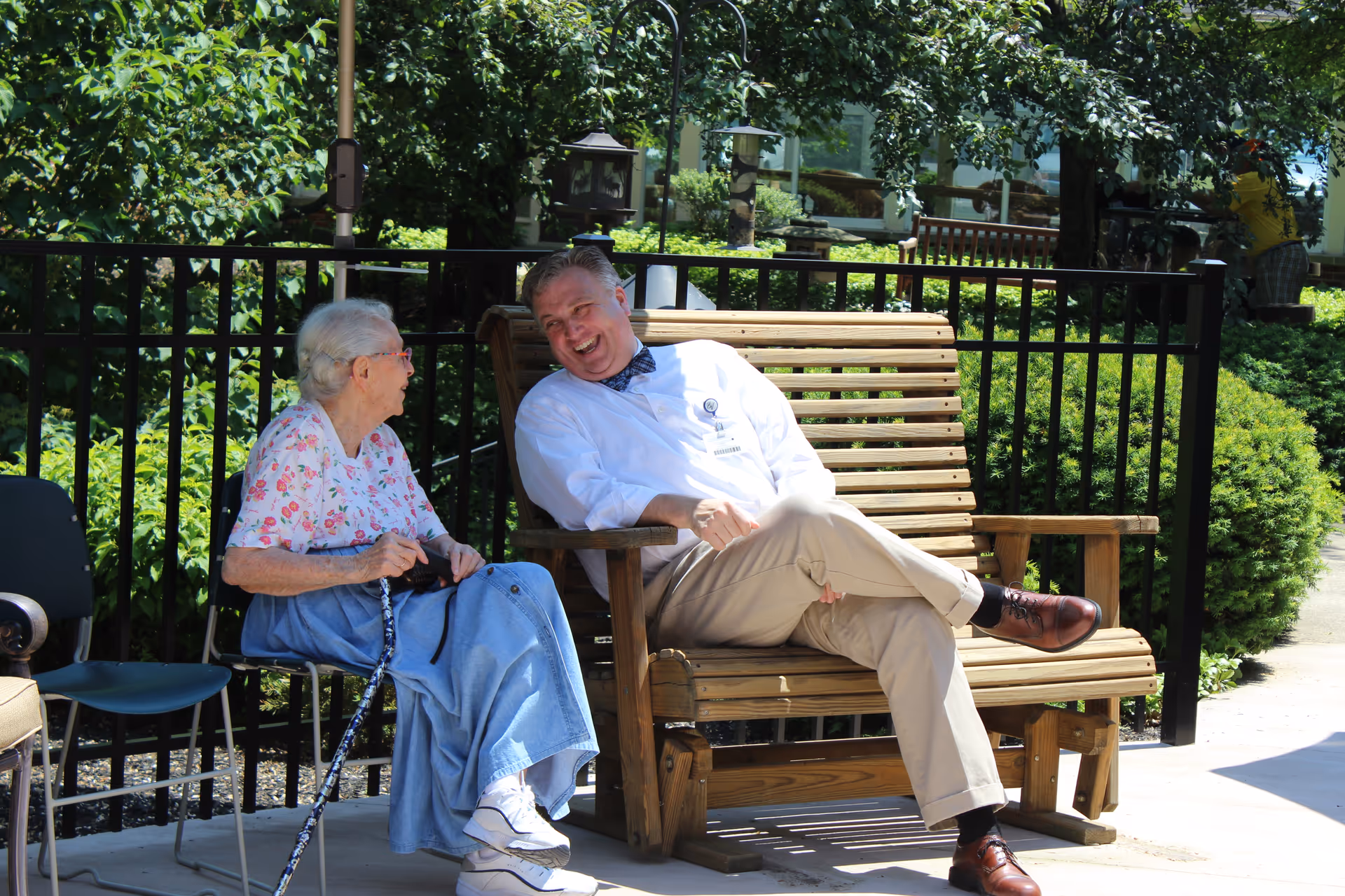 An elderly woman and a man in a white coat sitting and talking on a wooden bench outdoors near a black metal fence with greenery in the background.