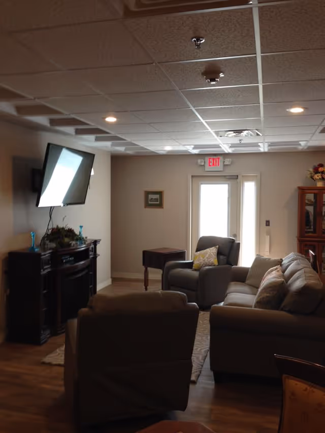 A cozy living room area with a mounted flat-screen TV above a dark wooden fireplace mantel. The room has a beige armchair, a gray armchair with a yellow patterned pillow, and a gray sofa with multiple cushions. There is a small wooden side table next to the gray armchair and a glass-front wooden cabinet with flowers on top. The room has a wooden floor, a patterned rug, and a door with a window letting in natural light. The ceiling has recessed lighting and a drop ceiling with tiles.
