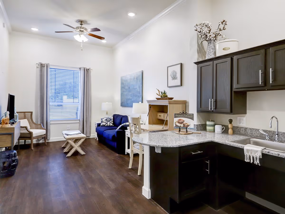 Interior view of a senior living facility room featuring a small kitchen area with dark cabinets and a granite countertop, a sink with a towel, and a living space with a blue couch, a wooden chair, a small table, a TV on a stand, and wall art. The room has wooden flooring, a ceiling fan with lights, and a window with blinds and curtains.