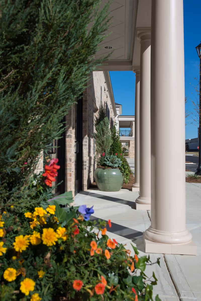 Outdoor walkway at The Blake at Flowood with large beige columns, brick walls, potted plants, and colorful flowers in the foreground under a clear blue sky.