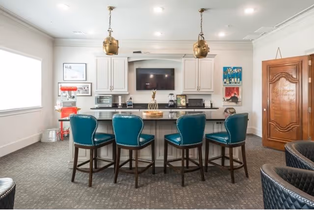 A modern kitchen area with a large island countertop surrounded by four teal upholstered bar stools. The kitchen features white cabinetry, a mounted flat-screen TV, and two brass pendant lights hanging from the ceiling. There is a popcorn machine on the left side near a window, and a wooden door on the right side with decorative carvings. The room is well-lit with recessed ceiling lights.