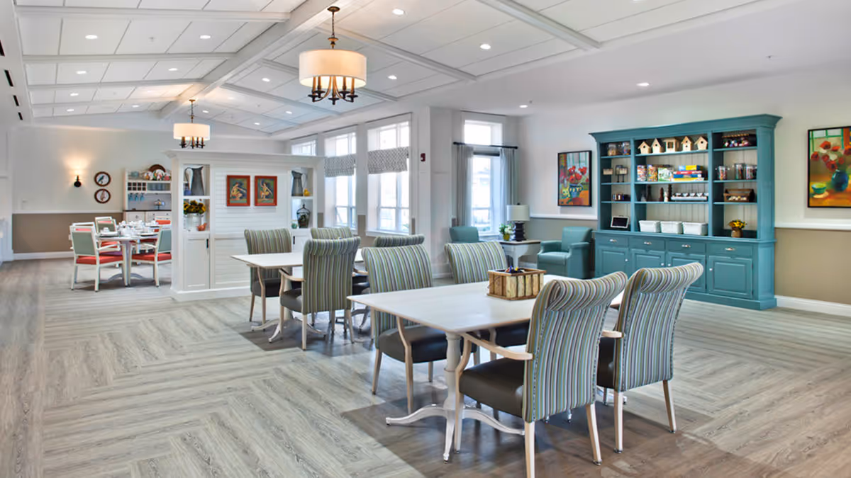 A bright and spacious dining area in a senior living facility with multiple tables and chairs. The chairs have striped upholstery, and the tables are white. There is a teal-colored cabinet filled with decorative items and games against one wall. The room has large windows with natural light, modern ceiling lights, and colorful artwork on the walls.