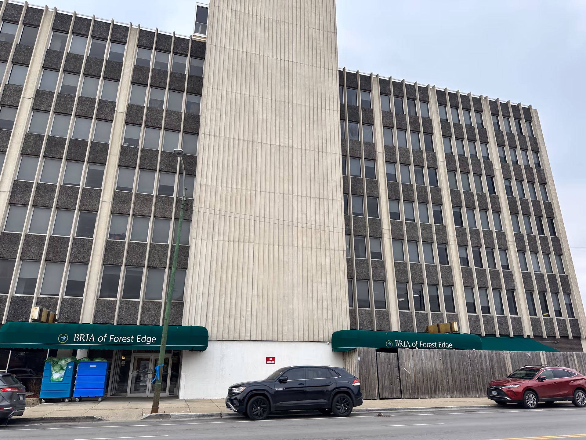 Exterior view of a multi-story building with green awnings displaying the name BRIA of Forest Edge. There are several windows on the building, two cars parked on the street in front, and a sidewalk with a street lamp and some blue recycling bins.