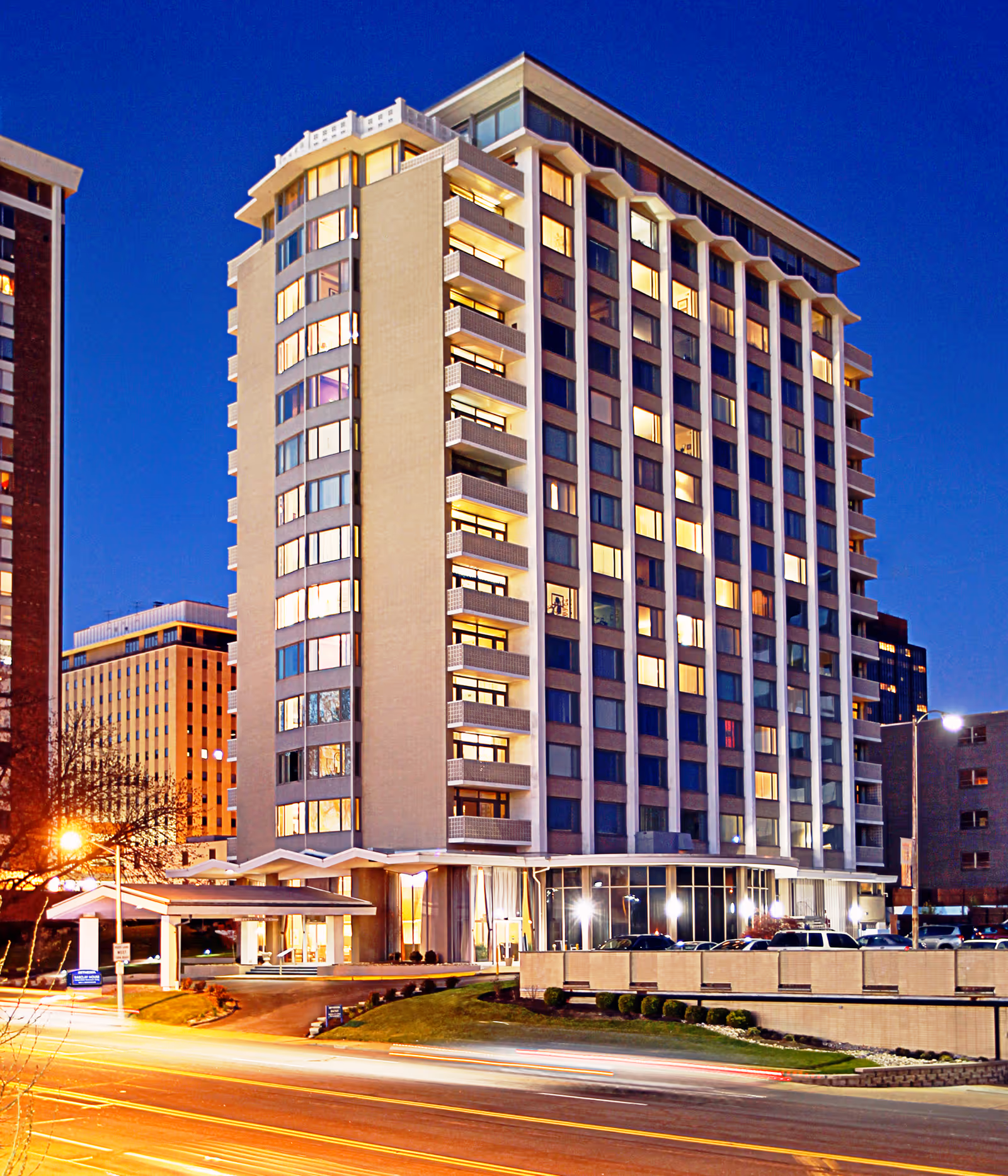 Tall multi-story residential building with illuminated windows and a covered entrance at dusk.
