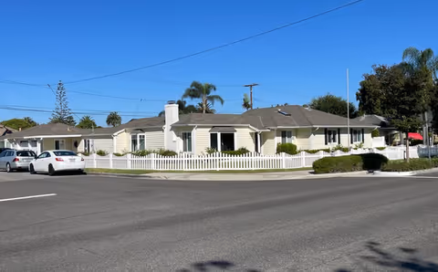 A single-story senior living facility with a white picket fence, parked cars, and palm trees.