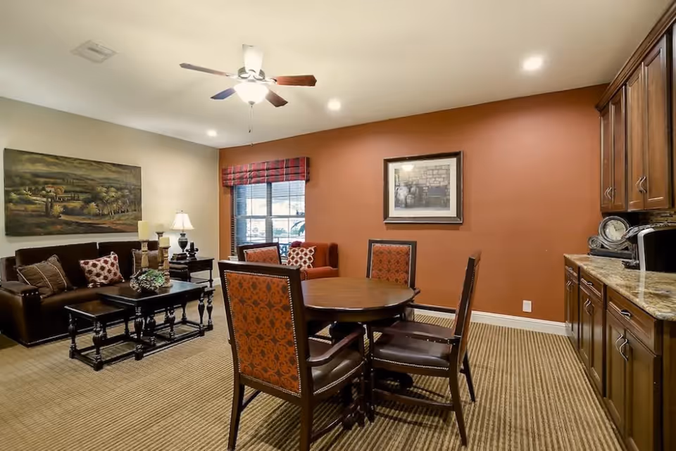 Well-lit communal living room with a sofa and coffee table, a round dining table with chairs, and cabinetry along the right wall.