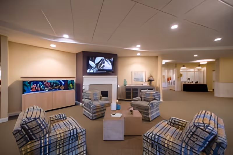 Spacious senior living lounge with patterned armchairs around a coffee table, a fireplace with a TV above, and a large aquarium along the back wall.
