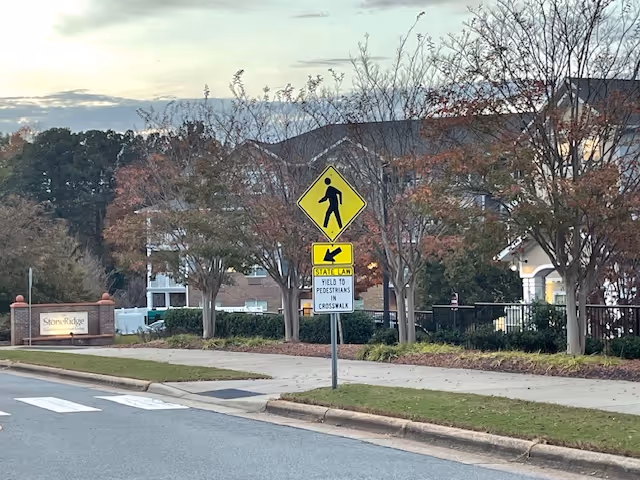 Street view showing a pedestrian crossing sign and a crosswalk in front of a sidewalk with trees and a building in the background. A sign for Stoneridge Gracious Retirement Living is visible on the left side near the sidewalk.