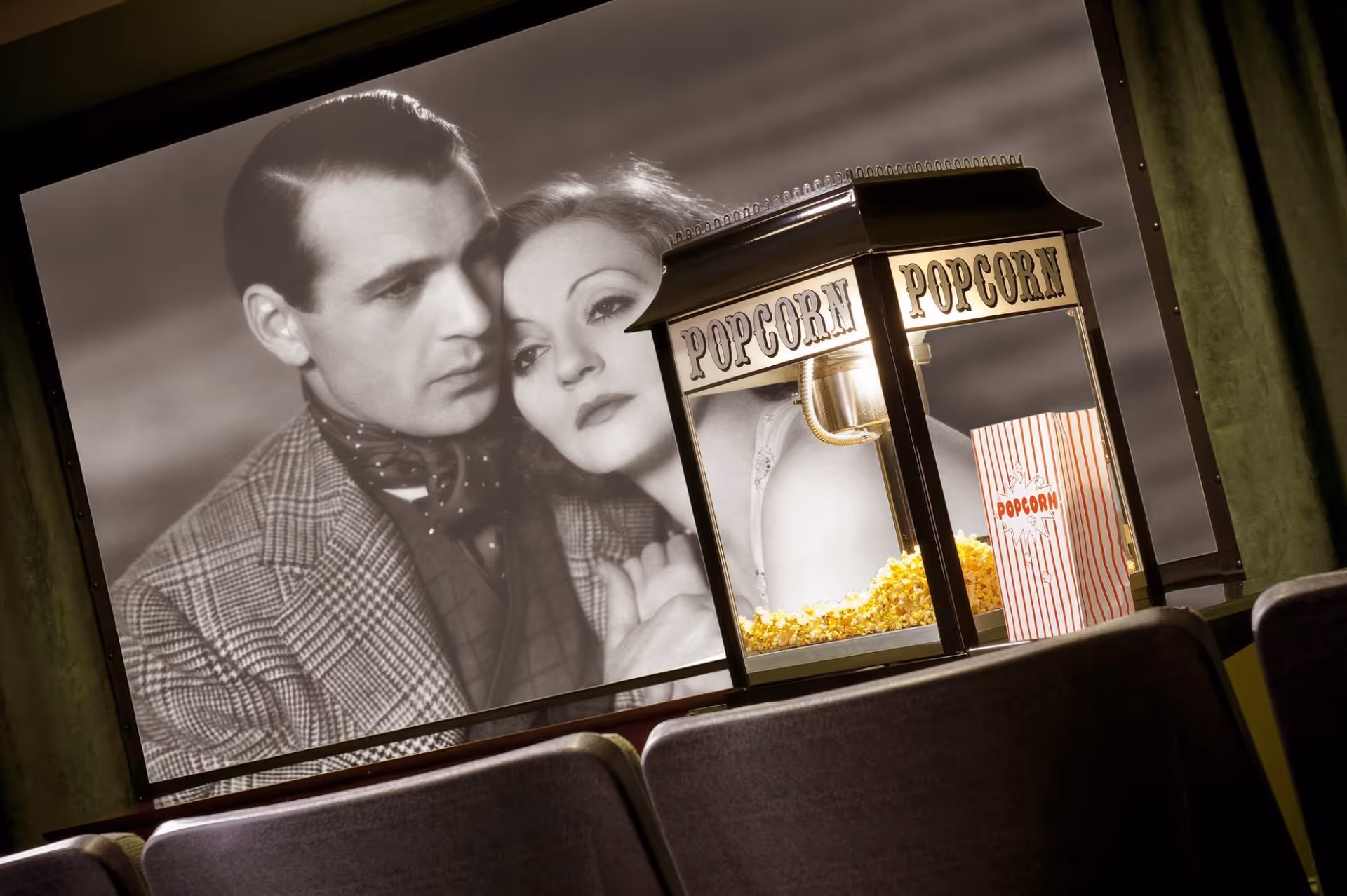 A vintage-style movie theater scene with a black and white film showing a man and woman in classic attire on the screen. In front of the screen, there is a popcorn machine filled with popcorn and a striped popcorn container. Theater seats are visible in the foreground.