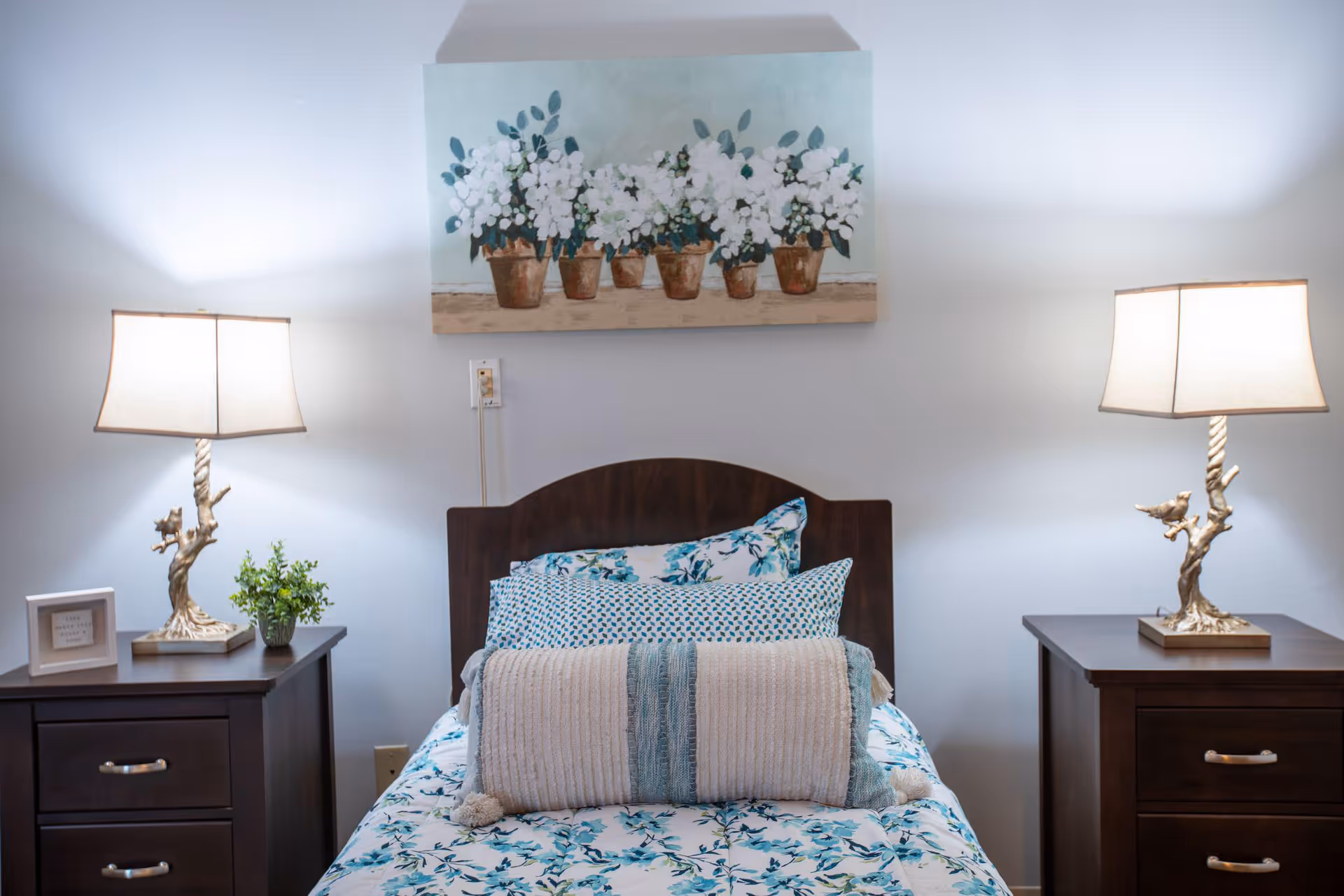 A neatly made single bed with floral bedding and multiple pillows is centered between two dark wooden nightstands, each with a decorative lamp and a small plant or framed picture. Above the bed hangs a painting of white flowers in brown pots against a light blue background.