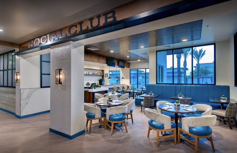 Interior view of a modern dining area named The Social Club with round tables, cushioned chairs with blue seats, a bar counter with stools, large windows showing palm trees outside, and contemporary lighting fixtures.