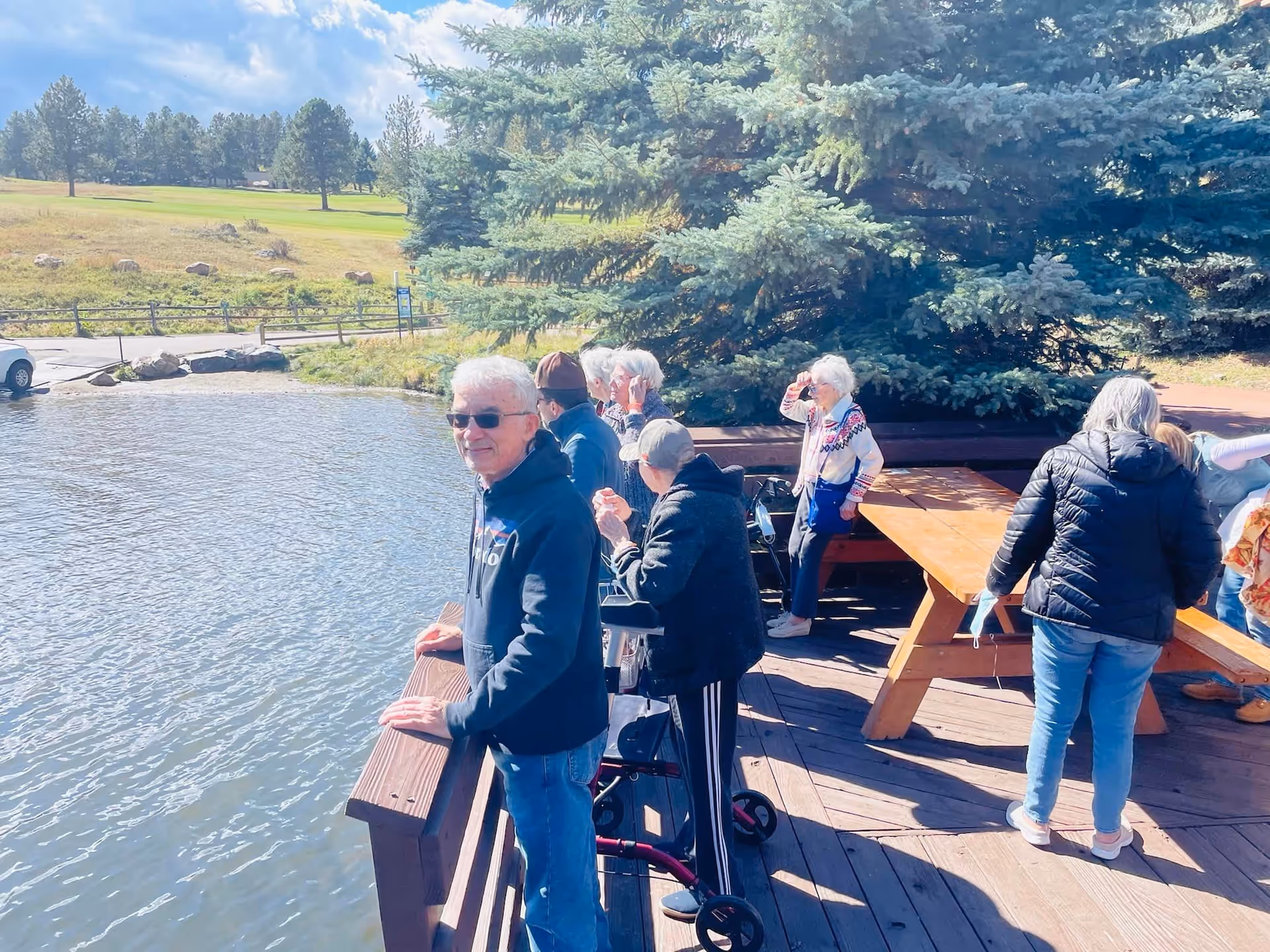 A group of elderly people standing and sitting on a wooden deck by a pond, surrounded by trees and greenery under a partly cloudy sky. Some individuals are looking at the water, while others are engaged in conversation or looking around.