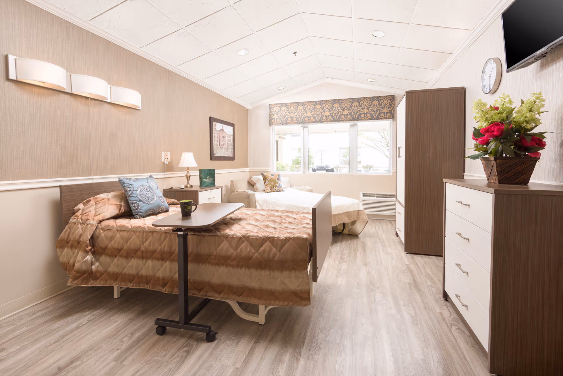 A bright and clean bedroom in a senior living facility featuring a single bed with a brown quilted bedspread and a blue decorative pillow. There is a small overbed table with a black mug on it. Next to the bed is a nightstand with a lamp and a green book. In the background, there is a white armchair with a floral pillow, a large window with patterned valance, a wooden wardrobe, and a dresser topped with a flower arrangement. A wall clock and a mounted TV are visible on the right wall. The room has light wood flooring and beige walls with white trim.