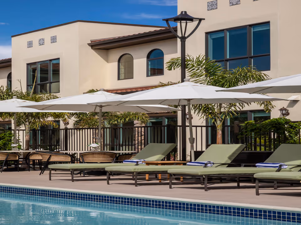 Outdoor pool area with several green cushioned lounge chairs and large white umbrellas. Behind the pool area is a beige building with large windows and some palm trees providing greenery. The sky is clear and blue.