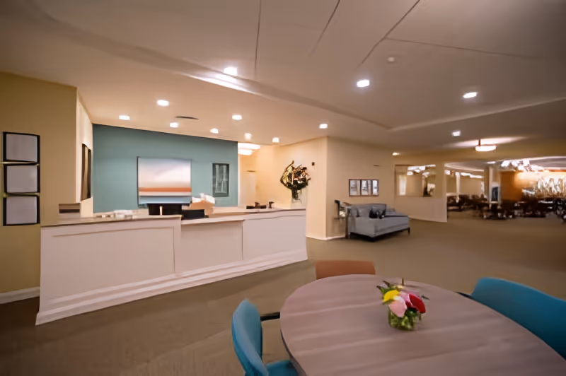 Interior view of a senior living facility reception area with a white front desk, a round table with colorful chairs and a small flower arrangement, a blue accent wall with artwork, and a seating area with a gray couch. The space is well-lit with ceiling lights and has a warm, welcoming atmosphere.
