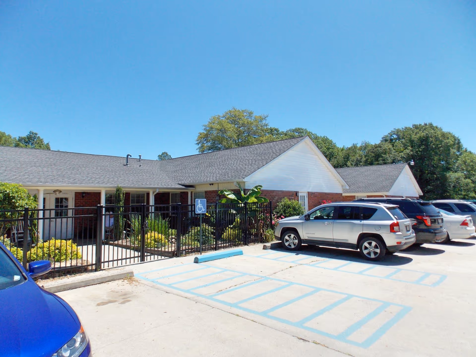Exterior view of Heath Springs Residential Care facility showing a single-story brick building with a gray shingled roof, a black metal fence enclosing a small garden area, several parked cars including a silver SUV and a blue car, and a clear blue sky above.