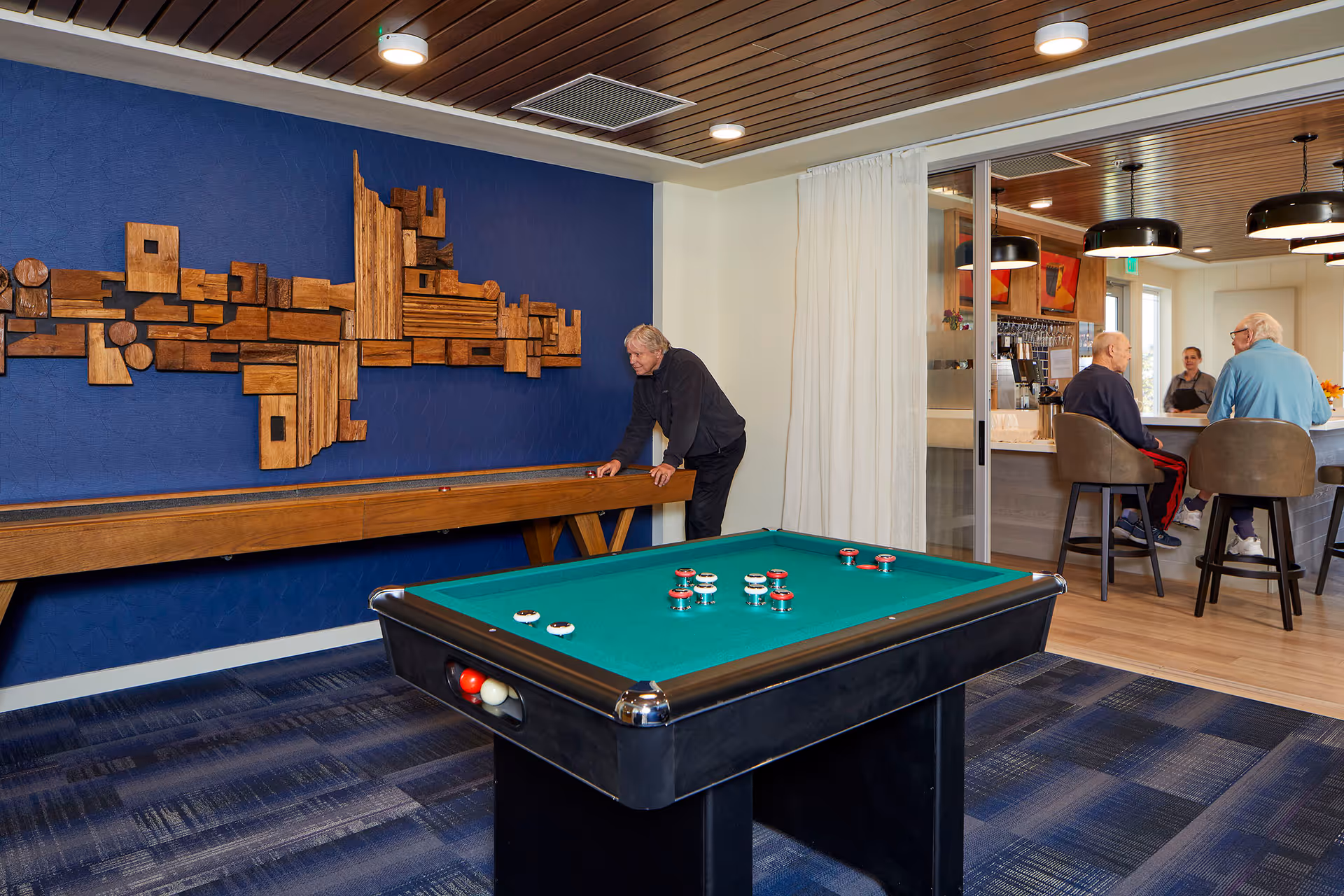 A recreational room in Revel Spokane featuring a shuffleboard table against a blue wall with wooden art, a billiards table in the foreground, and a bar area in the background where two elderly men sit on stools talking to a bartender.