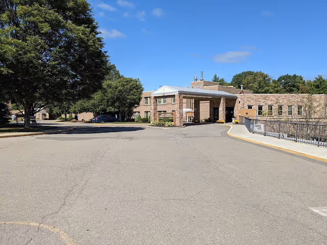 Exterior view of a brick building with a covered entrance driveway, surrounded by trees and a clear blue sky.