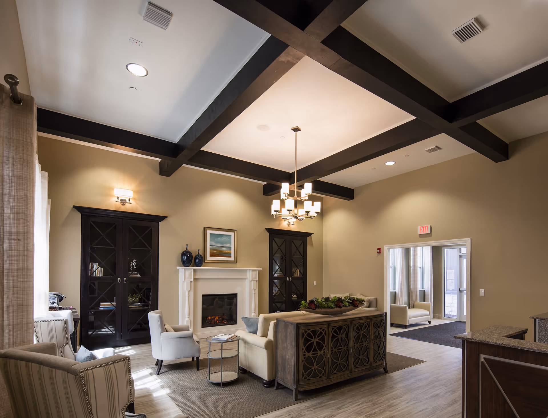 A cozy living room area in a memory care facility with beige walls and dark wooden ceiling beams. The room features a white fireplace with a painting above it, two black glass-door cabinets on either side, a beige sofa, two armchairs, a small round table, and a wooden console table with decorative plants. There is a modern chandelier hanging from the ceiling and an exit door leading to another room with more seating.
