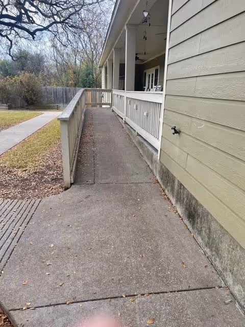 A concrete wheelchair ramp with handrails on the left side leading up to a covered porch area of a building with light green siding. There are trees and a grassy area visible in the background.