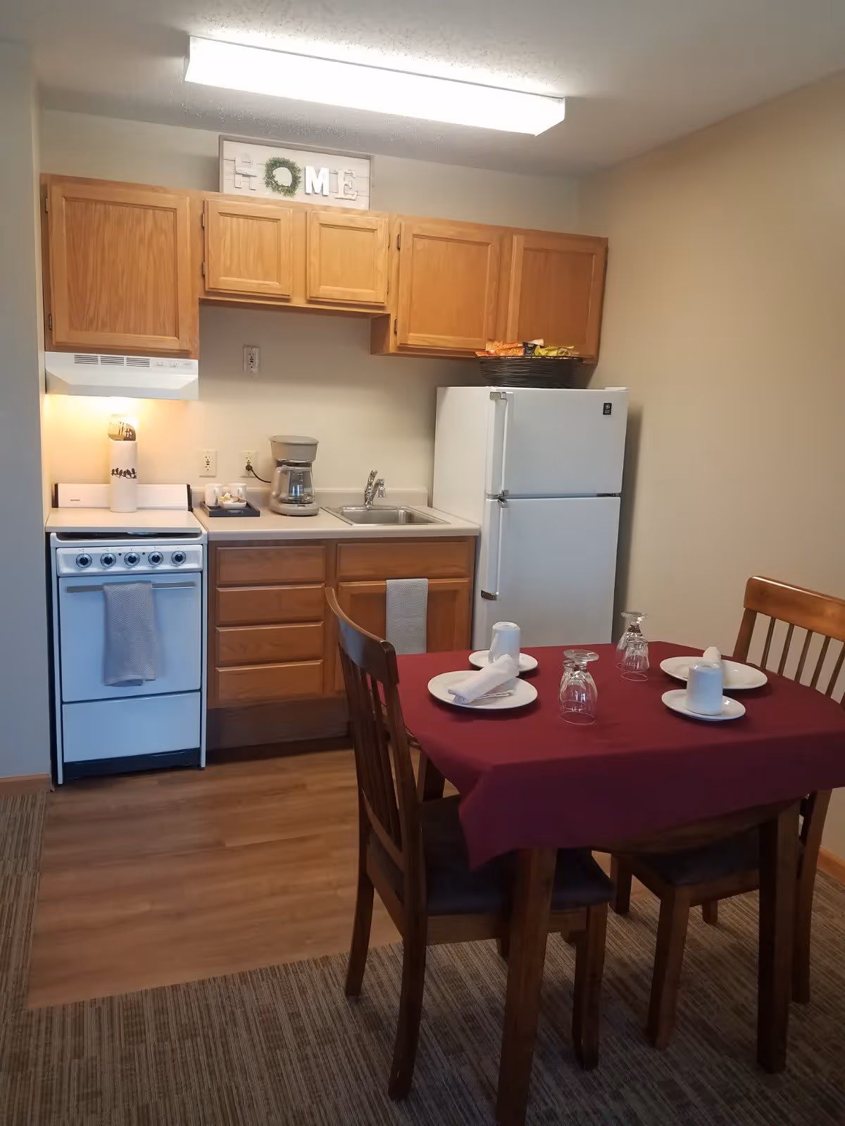 A small kitchen area with wooden cabinets, a white stove with a towel hanging on the handle, a coffee maker on the counter, a sink, and a white refrigerator. In front of the kitchen is a dining table covered with a burgundy tablecloth, set with four plates, napkins, and upside-down glasses. The floor is a combination of wood and carpet, and there is a decorative sign above the cabinets that spells 'HOME' with a wreath as the letter O.
