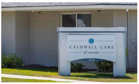 Outdoor view of a building wall with a sign in front that reads 'Caldwell Care of Cascadia' with the number 210 below it, surrounded by grass and small bushes.