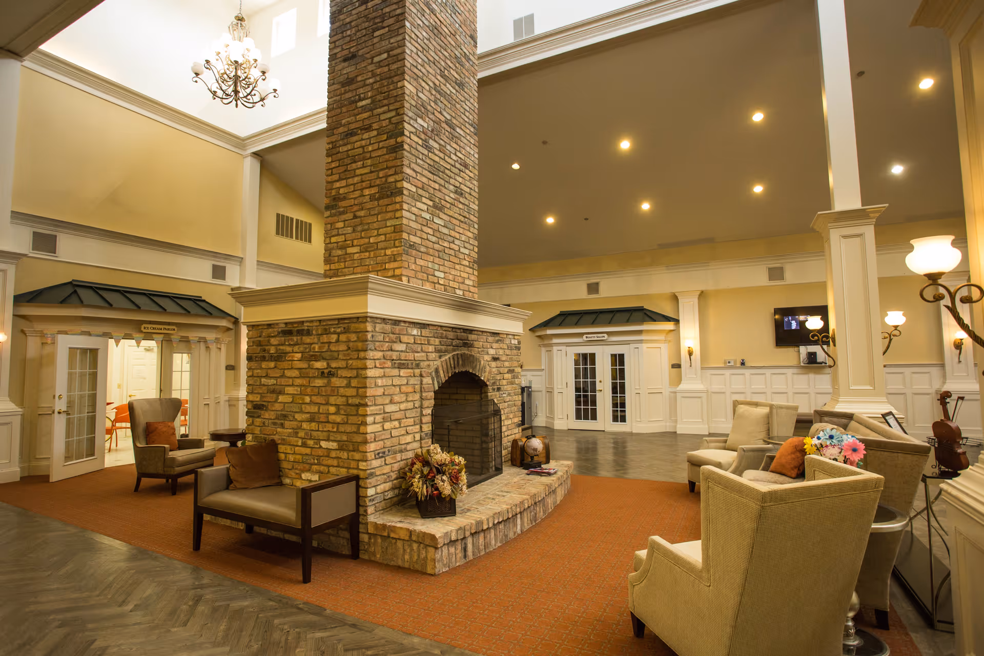 A spacious and warmly lit living room area in a senior living facility featuring a large brick fireplace in the center. The room has comfortable armchairs and sofas arranged around the fireplace, decorative lamps, and a chandelier hanging from the high ceiling. There are two doorways with small awnings labeled 'Ice Cream Parlor' and 'Beauty Salon'.