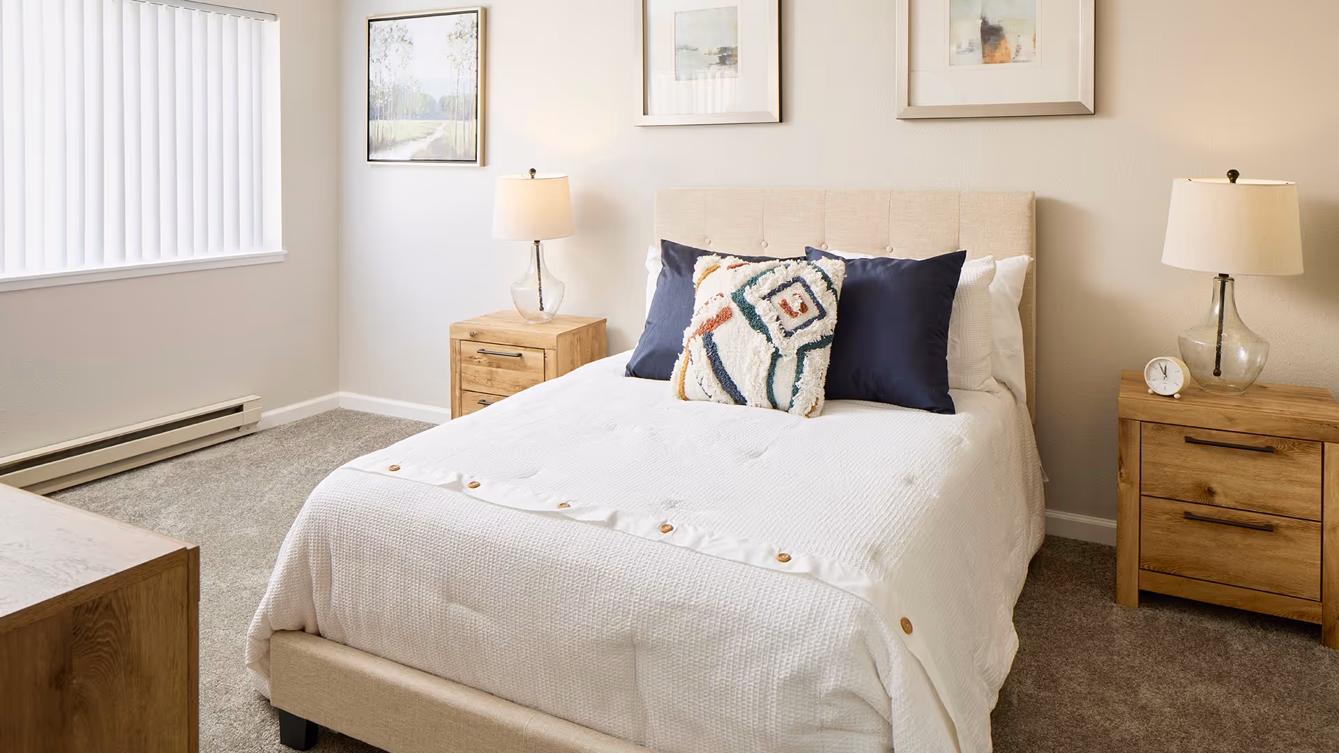 A bright bedroom with a beige upholstered bed featuring white bedding and decorative pillows. There are two wooden nightstands on either side of the bed, each with a glass lamp and one with a small clock. The walls are light-colored with three framed artworks above the bed. A window with vertical blinds is on the left side of the room.