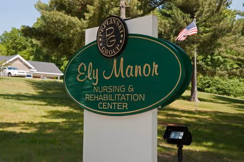 Outdoor green sign reading Ely Manor Nursing and Rehabilitation Center amid trees and a flag.