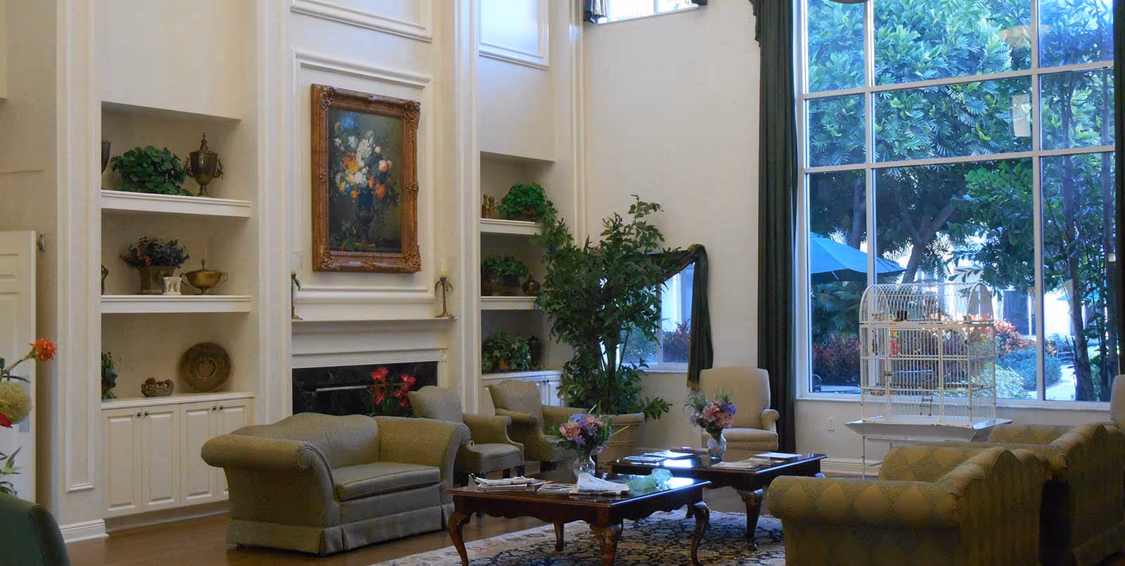 A spacious and elegant living room with high ceilings, featuring comfortable upholstered sofas and armchairs arranged around two wooden coffee tables with floral centerpieces. The room has built-in white shelves with decorative items, a large framed floral painting above a fireplace, and a large window with green curtains overlooking lush greenery outside. A white birdcage is also visible near the window.