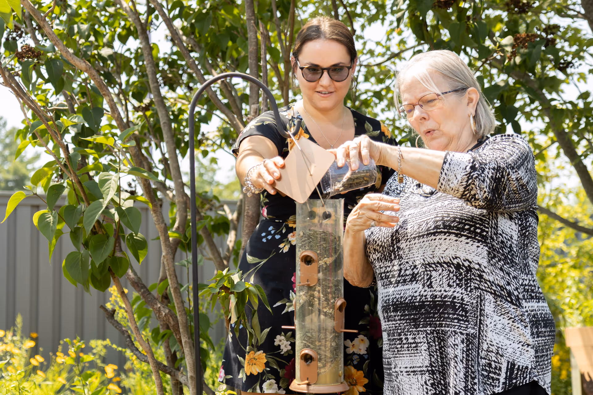 An elderly woman and a younger woman are outdoors in a garden setting. The elderly woman is pouring birdseed into a transparent bird feeder while the younger woman watches and assists. They are surrounded by green trees and plants, and the scene is bright and sunny.