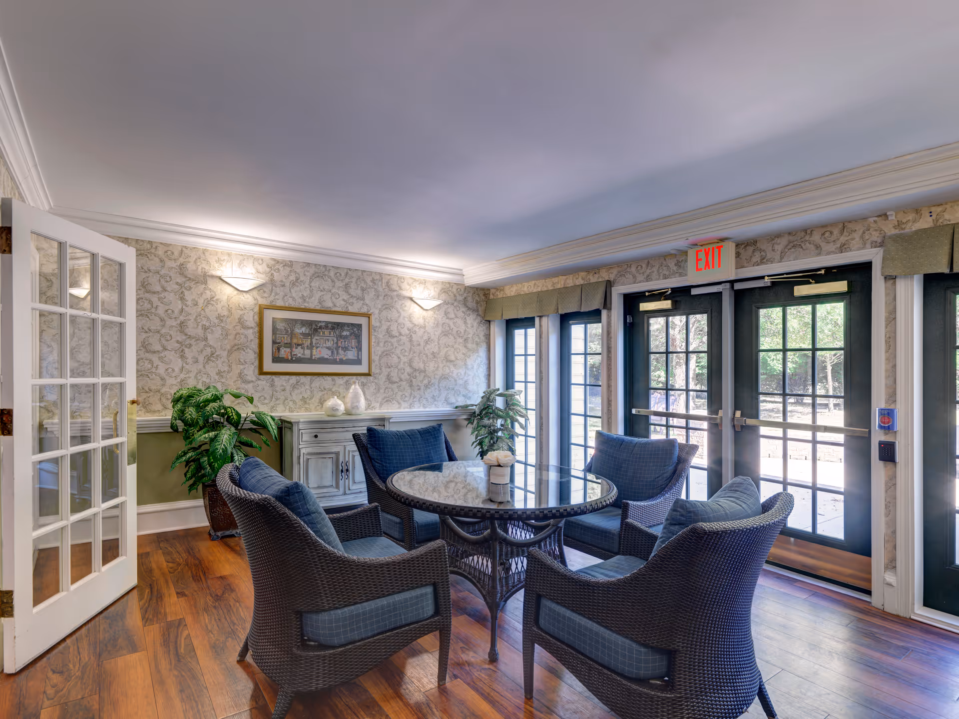 A cozy sitting area with four wicker chairs with blue cushions arranged around a round glass-top table. The room has wooden flooring, patterned wallpaper, and a sideboard with decorative items. There are large glass doors and windows letting in natural light, and an exit sign above the doors.