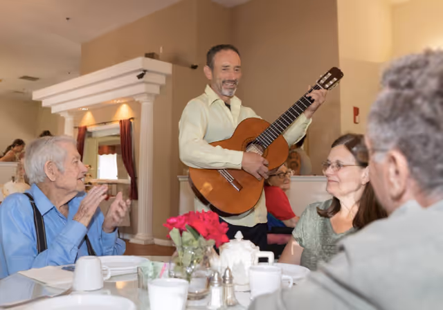 A man playing an acoustic guitar and smiling while standing in a dining area of a senior living facility. Several elderly people are seated around a table with white dishes, cups, and a vase with red flowers, enjoying the music. The room has beige walls and decorative columns.