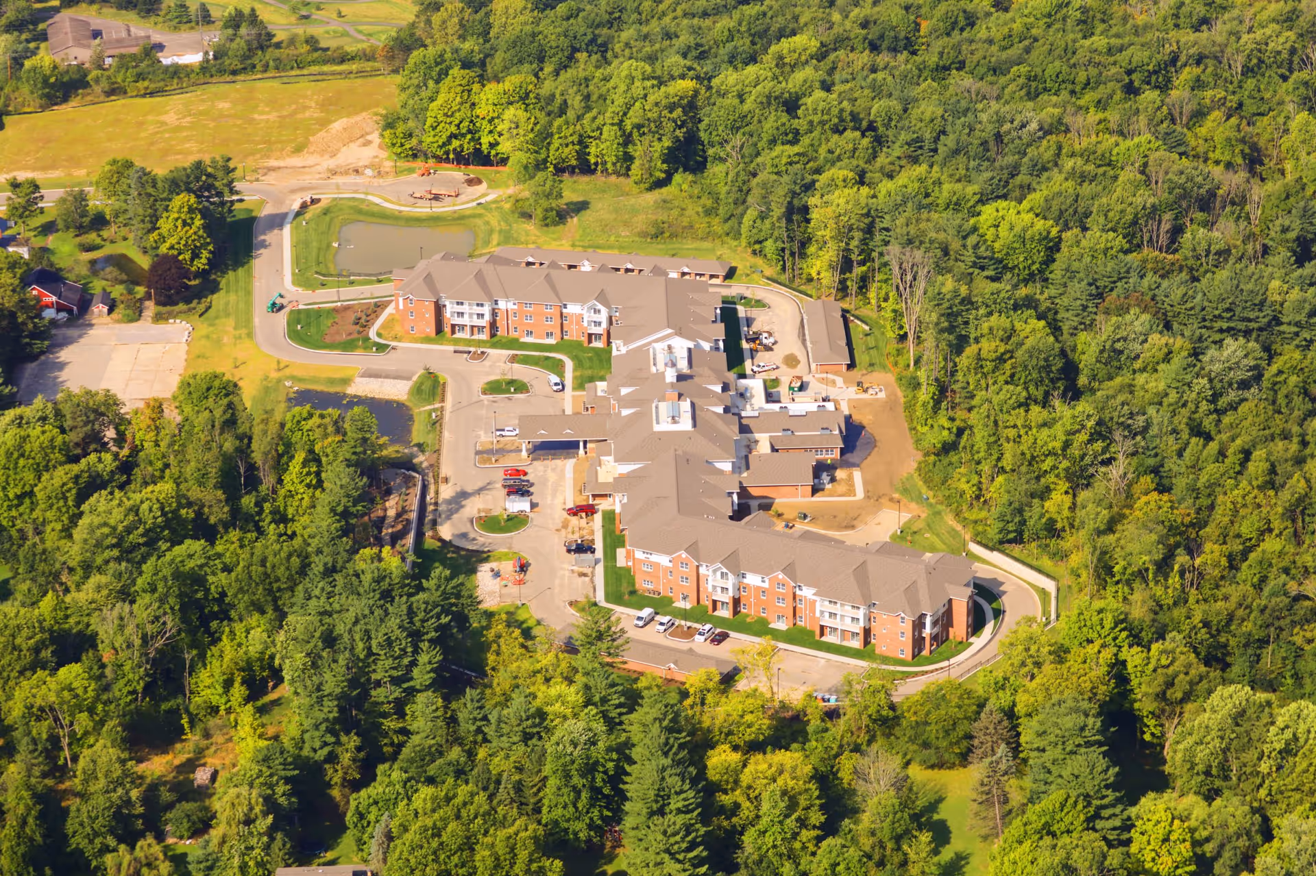 Aerial view of Hudson Meadows Retirement Community surrounded by dense green trees and vegetation, showing a large multi-wing building with parking areas and a small pond nearby.