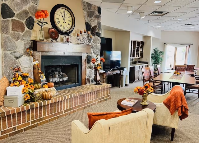A cozy living room area with a stone and brick fireplace decorated with autumn-themed items including pumpkins, flowers, and a clock above the mantel. Two beige armchairs with orange pillows and a throw blanket face a small round table with a floral centerpiece. In the background, there is a TV on a stand, a dining table with chairs, and large windows letting in natural light.