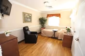 A small, clean room with a single bed covered in a beige bedspread, a black recliner chair, a wooden dresser, a small table on wheels, a wall-mounted TV, a framed picture, and a window with a curtain. The floor is wooden, and there is a potted plant in the corner.